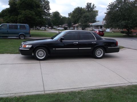 2001 Mercury Grand Marquis - A black 2001 Mercury Grand Marquis sedan is parked on a concrete driveway in front of a residential house. The car is viewed from the side,…