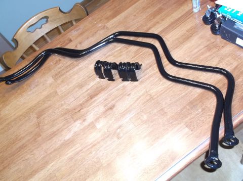1999 Mercury Grand Marquis - Glossy black sway bar with its mounting brackets and bushings arranged on a light wood table, chair behind.