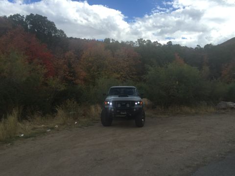 1988 Ford Bronco - A lifted, dark-colored SUV is parked on a dirt road with a backdrop of trees showing fall colors. The vehicle has its headlights on and app…