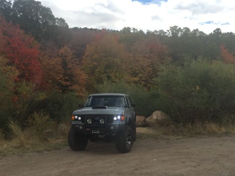 1988 Ford Bronco - A lifted, gray 1988 Ford Bronco is parked on a dirt road in front of a hillside covered in autumn foliage. The vehicle has a black bull bar…