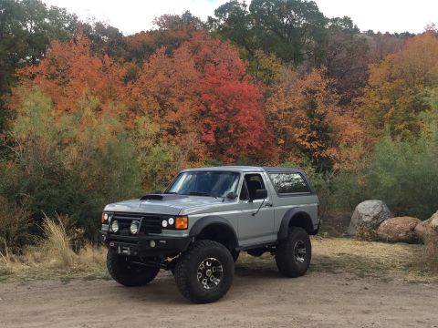 1988 Ford Bronco - A lifted, silver 1988 Ford Bronco is parked on a dirt road with a backdrop of trees displaying vibrant fall foliage. The vehicle features a…