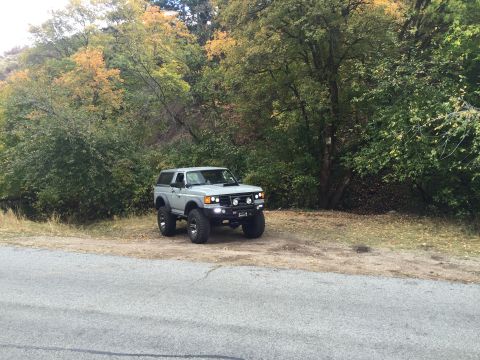 1988 Ford Bronco - A lifted, light gray SUV is parked off-road next to a paved road, with a backdrop of trees displaying autumn foliage.