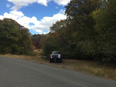 1988 Ford Bronco - A dark colored SUV is parked on the side of a paved road next to a dense forest with fall foliage. The vehicle's headlights are illuminated.