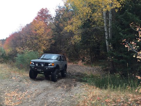 1988 Ford Bronco - A grey Ford Bronco is parked on a dirt road surrounded by trees with autumn foliage. The vehicle has a lifted suspension and an aftermarket…
