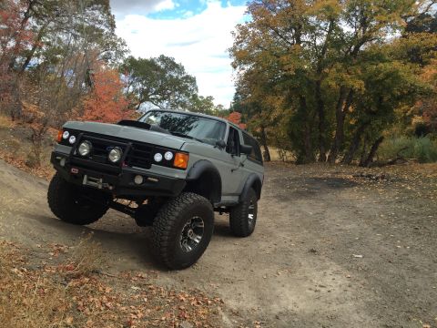 1988 Ford Bronco - A lifted gray Ford Bronco is parked on a dirt road with autumn foliage in the background. The off-road vehicle features a custom bumper wit…