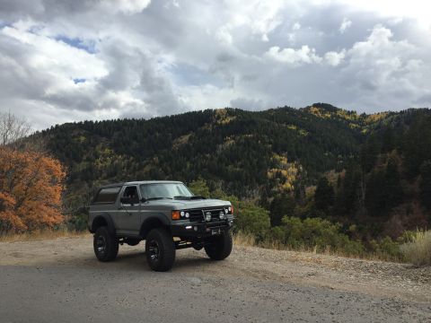 1988 Ford Bronco - A gray Ford Bronco is parked on a dirt road with a wooded mountain landscape in the background. The vehicle has off-road modifications incl…