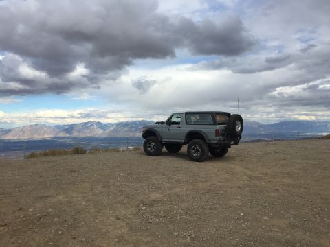 1988 Ford Bronco - A lifted, two-door 1988 Ford Bronco is parked on a dirt overlook with a scenic mountain range in the background under a cloudy sky.