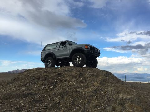 1988 Ford Bronco - A lifted, gray 1988 Ford Bronco is parked on a rocky hill under a cloudy sky. The vehicle has large off-road tires and an aftermarket bumpe…