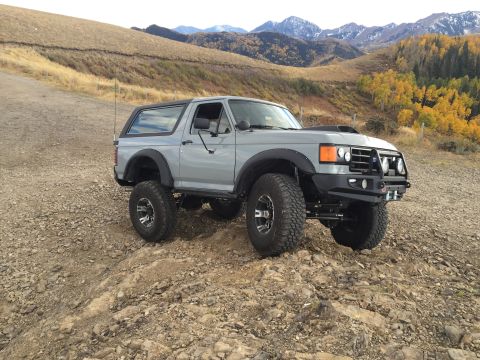 1988 Ford Bronco - A lifted, gray 1988 Ford Bronco is parked on a rocky, off-road trail with mountains and autumn foliage in the background.