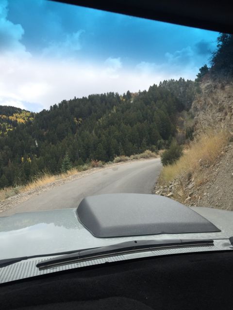 1988 Ford Bronco - View from inside a vehicle driving on a winding paved road through a forested mountain landscape. The hood of the vehicle, with a dark gray…