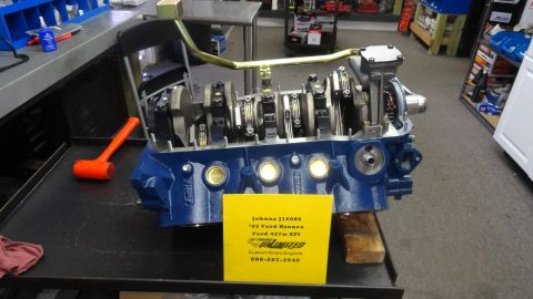 1982 Ford Bronco - A blue Ford 427w EFI engine is displayed on a workbench with a yellow sign detailing its specifications. A rubber mallet rests to the left…