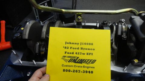1982 Ford Bronco - A yellow card with engine details is held in front of a Ford 427w EFI engine, likely from a 1982 Ford Bronco. The card lists "Johnny J16086…