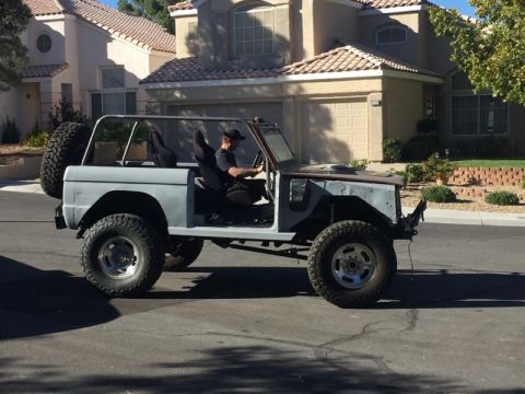 1979 Ford Bronco - person drives a primer-gray early-model Ford Bronco with the top removed and oversized tires past stucco houses.