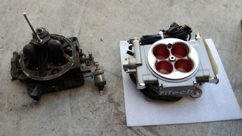 1979 Ford Bronco - Two fuel injection throttle bodies are shown side-by-side on a concrete surface. The older, darker unit on the left is a carburetor, while…