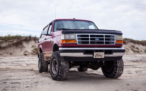 1995 Ford Bronco - A lifted, maroon 1995 Ford Bronco is parked on a sandy beach with dunes and sparse vegetation in the background. The front of the SUV is fa…