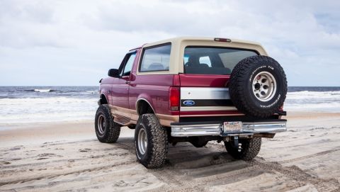 1995 Ford Bronco - The rear driver's side view of a maroon and tan 1995 Ford Bronco parked on a sandy beach with the ocean in the background. The SUV has a sp…