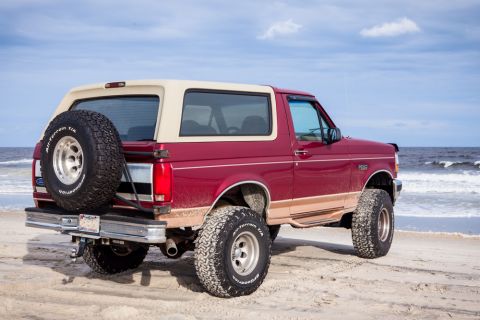 1995 Ford Bronco - A red 1995 Ford Bronco with a tan removable top is parked on a sandy beach with the ocean in the background. The vehicle has large off-road…