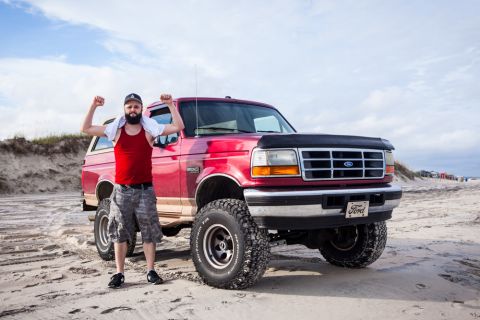 1995 Ford Bronco - A man wearing a red tank top and camouflage shorts flexes his arms in front of a red Ford Bronco on a sandy beach. The Bronco has large off…