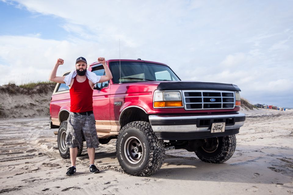 Man flexing in front of a red Ford Bronco on a beach.