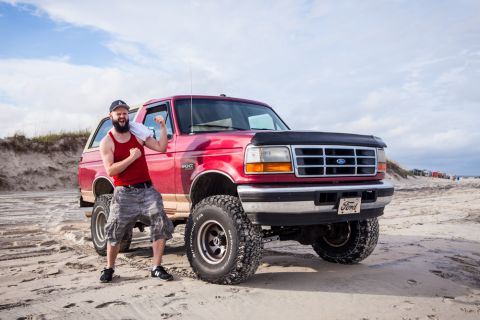 1995 Ford Bronco - A man in a red tank top and camouflage shorts poses triumphantly next to a lifted red Ford Bronco on a sandy beach. The Bronco has large of…