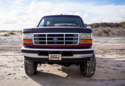 1995 Ford Bronco - Front view of a maroon Ford Bronco parked on a sandy beach with dunes in the background. The vehicle has a chrome bumper and grille with a…