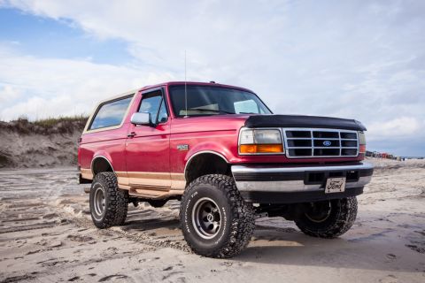 1995 Ford Bronco - A red 1995 Ford Bronco with tan trim sits on a sandy beach under a cloudy sky. The SUV has large off-road tires and a black bug deflector o…