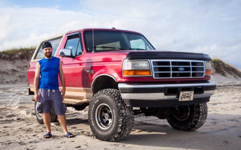 1995 Ford Bronco - A man stands next to a red 1990s Ford Bronco on a sandy beach with dunes in the background. The Bronco has large off-road tires and a Ford…