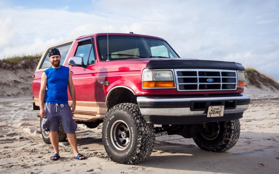 Man standing next to a red Ford Bronco on a sandy beach.