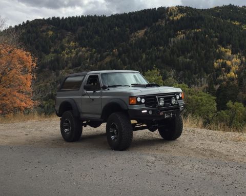 1988 Ford Bronco - A lifted, gray 1988 Ford Bronco is parked on a dirt road with a wooded mountainside in the background during autumn.