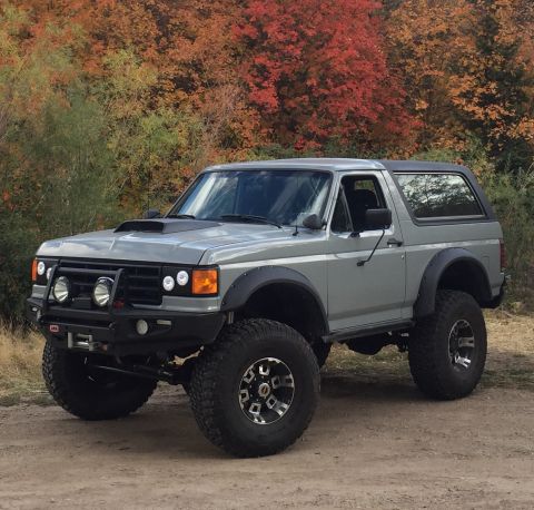 1988 Ford Bronco - A lifted, grey 1980s Ford Bronco with off-road tires and a custom bumper sits parked on a dirt road with colorful autumn trees in the backg…