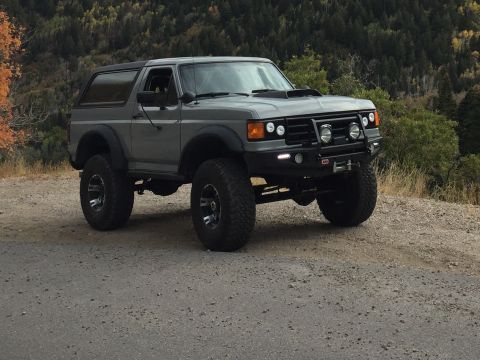 1988 Ford Bronco - A gray 1988 Ford Bronco with off-road modifications sits on a gravel road with a wooded hillside in the background. The vehicle features la…
