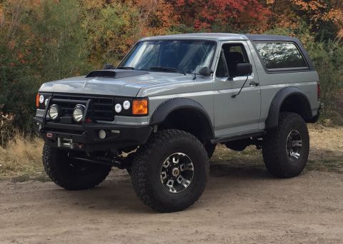 1988 Ford Bronco - A lifted, gray 1988 Ford Bronco with a black hood scoop and fender flares sits parked on a dirt road with colorful autumn foliage in the ba…
