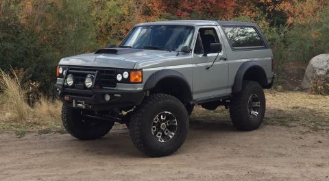 1988 Ford Bronco - A lifted, gray Ford Bronco with off-road tires and a custom bumper sits on a dirt road with autumn foliage in the background.
