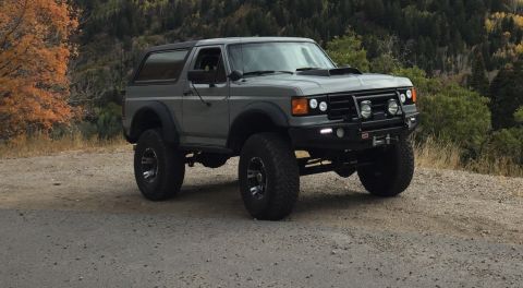 1988 Ford Bronco - A lifted, gray 1980s Ford Bronco is parked on a gravel road with a wooded hillside in the background.