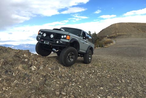 1988 Ford Bronco - A lifted, gray Ford Bronco is shown at an angle on a rocky incline with a mountainous landscape and cloudy sky in the background.