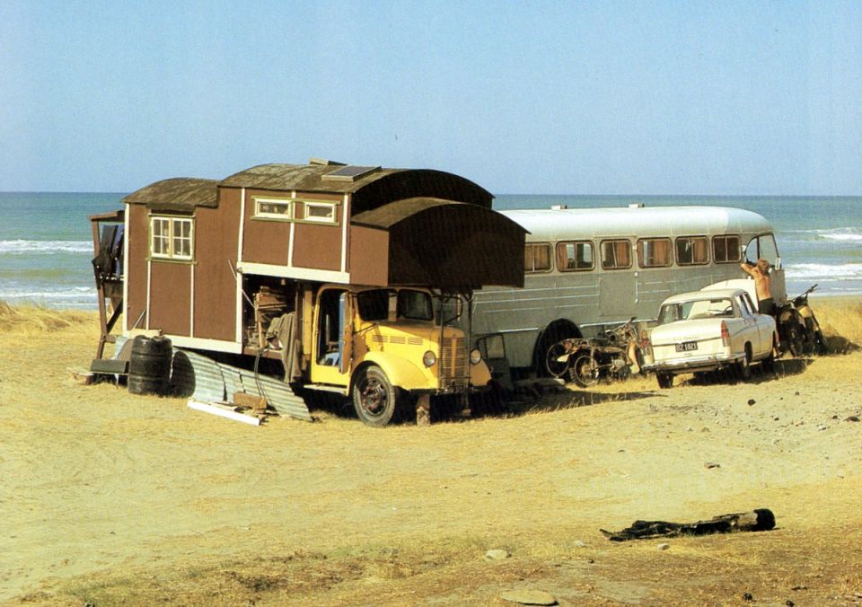 Modified camper on a truck chassis, vintage bus, and white sedan on a sandy beach.