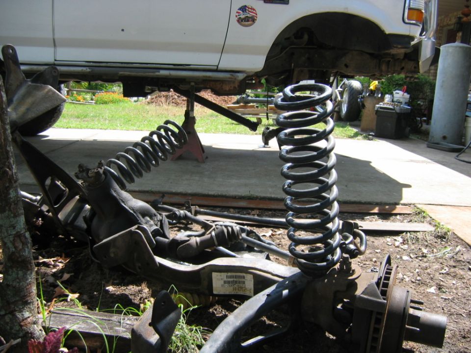 White Ford truck raised on jack stands, front suspension parts and coil spring on the ground.