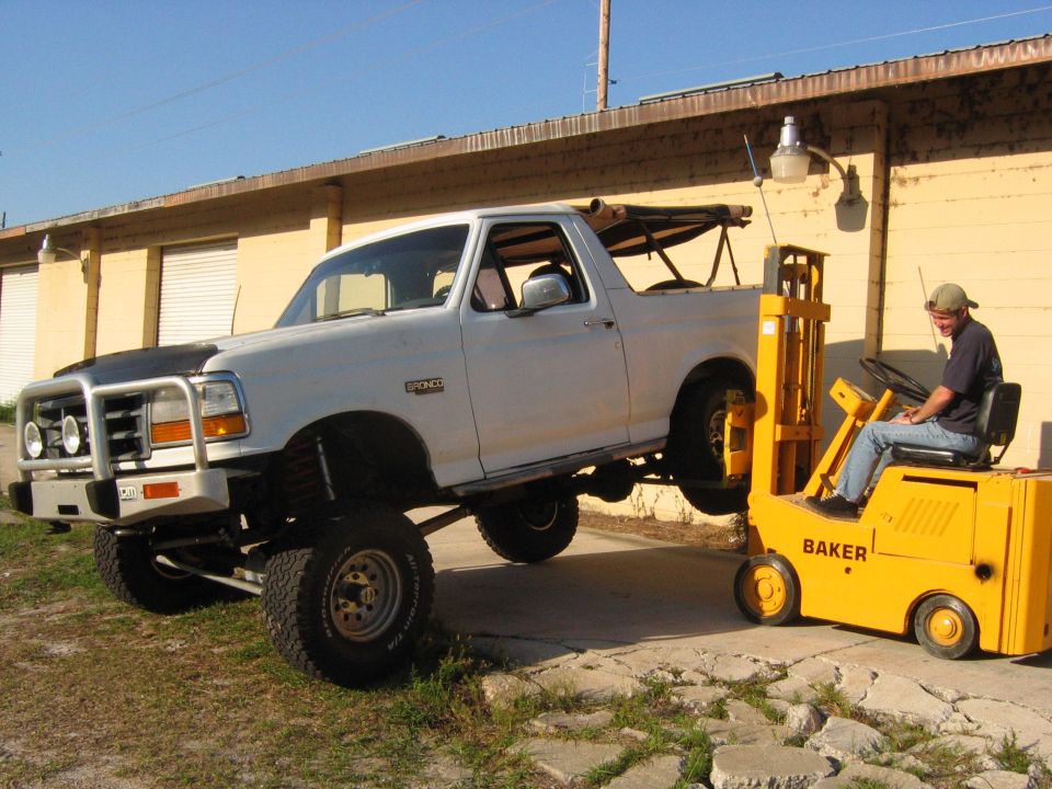 Lifted white Ford Bronco being moved by a yellow Baker forklift with a man driving.