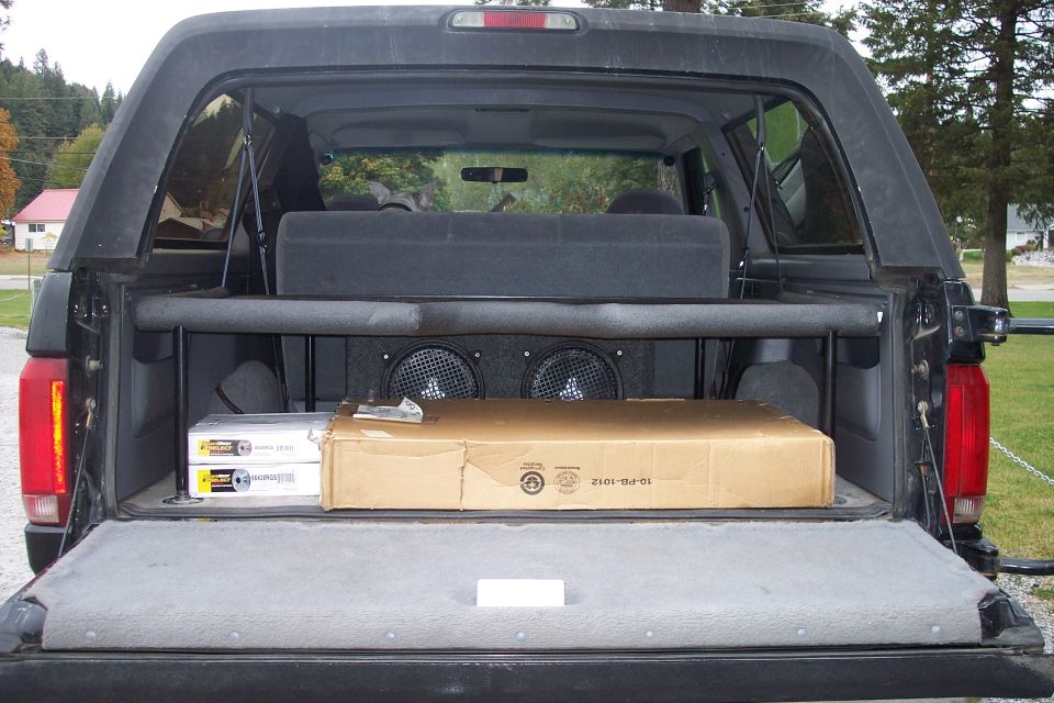 Open tailgate of a black Ford Bronco showing a subwoofer box, speakers, and boxes inside.