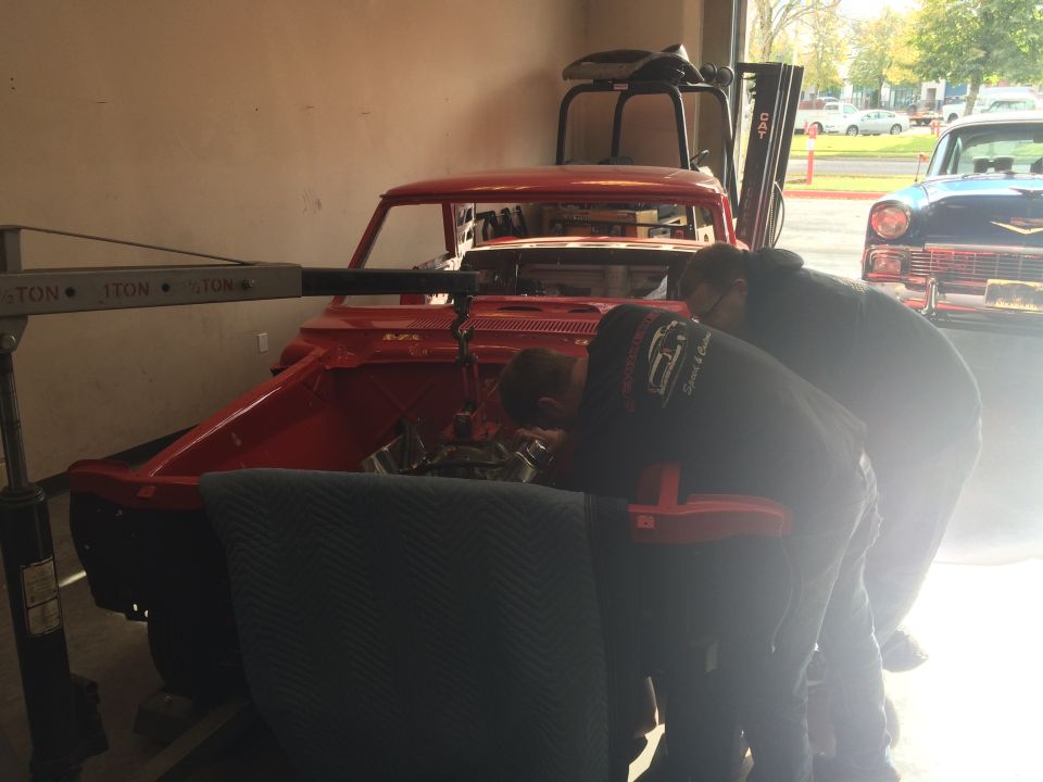 Red car body with engine removed, being worked on by two mechanics; blue 1957 Chevrolet visible to the right