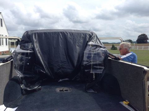 1979 Ford Bronco - A vehicle is covered by a black tarp in the bed of a pickup truck, with a man observing.