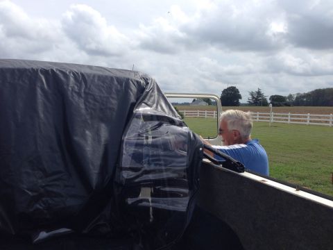 1979 Ford Bronco - A man sits in the driver's seat of a vehicle partially covered by a black tarp, with a rural landscape visible behind.