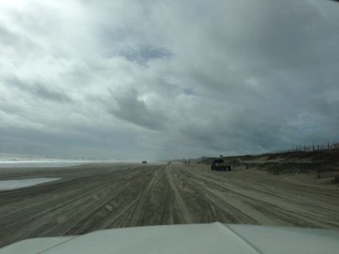 1979 Ford Bronco - Vehicles drive along the beach, leaving tire tracks in the sand under a dramatic sky.