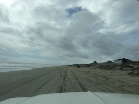 1979 Ford Bronco - Vehicles traverse a sandy path along the coast, with houses visible on a dune overlooking the beach.