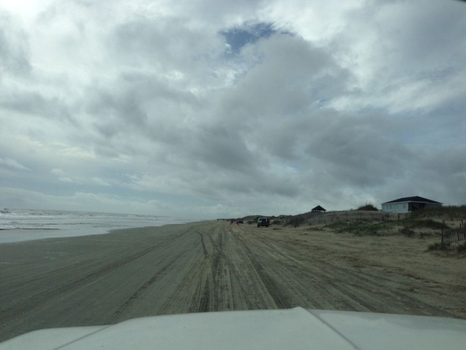 Several vehicles drive on a sandy road next to the ocean under a cloudy sky
