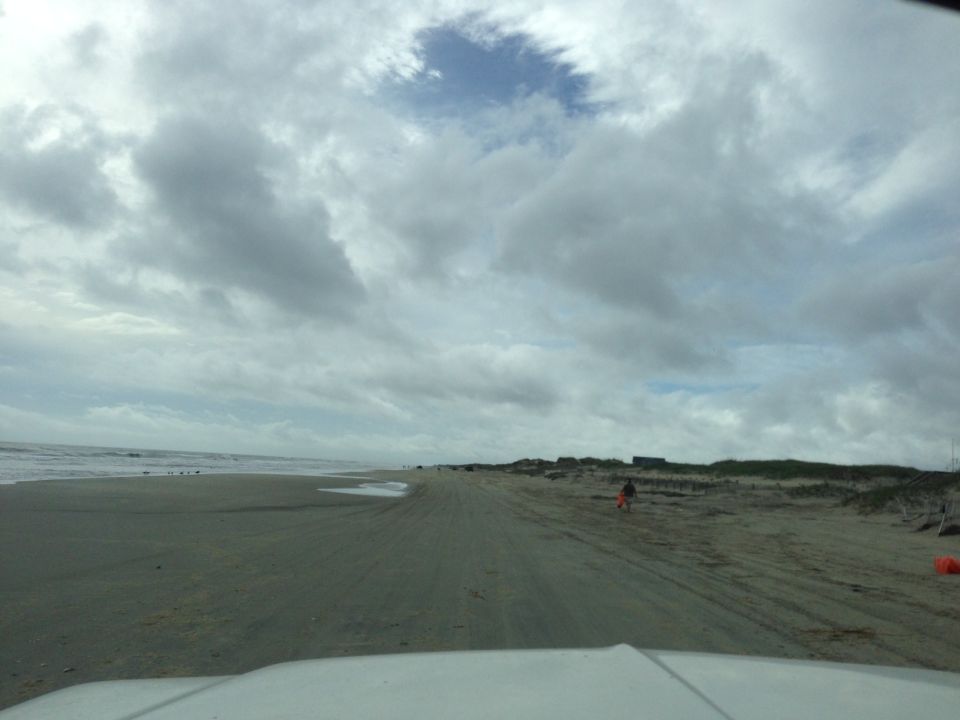 White vehicle hood in foreground, driving on a sandy beach with ocean waves and cloudy sky