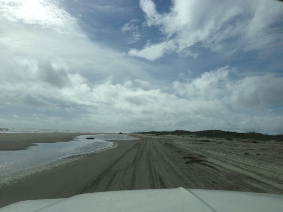 View from inside a vehicle driving on a sandy beach with tire tracks and ocean in the distance