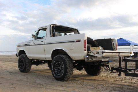 1979 Ford Bronco - A white, lifted Ford Bronco is parked on a sandy beach with its tailgate down. A blue canopy is visible in the background.