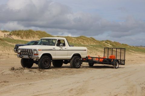 1979 Ford Bronco - A lifted white 1970s Ford pickup truck tows a utility trailer loaded with orange items on a sandy beach. The truck has large off-road tires…