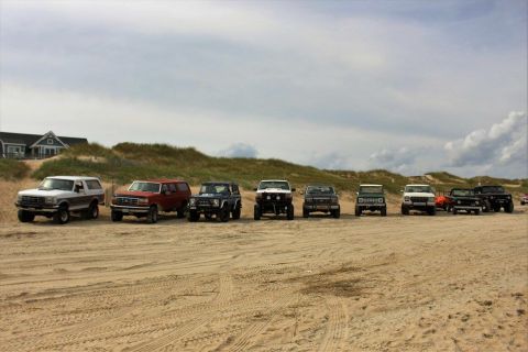 1979 Ford Bronco - A line of SUVs and trucks are parked on a sandy beach with dunes and a house in the background under a cloudy sky.